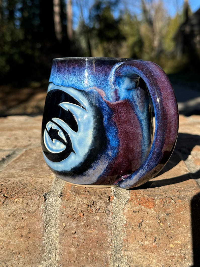 May include: A blue and purple ceramic mug with a black and white swirl design. The mug has a handle and is sitting on a brick surface.