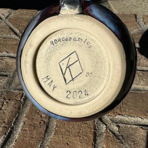 May include: A close-up of the base of a dark blue ceramic mug with a white glaze. The mug has a white stamp that reads "MAY 2024" and "404ceramics" with a logo in the centre.