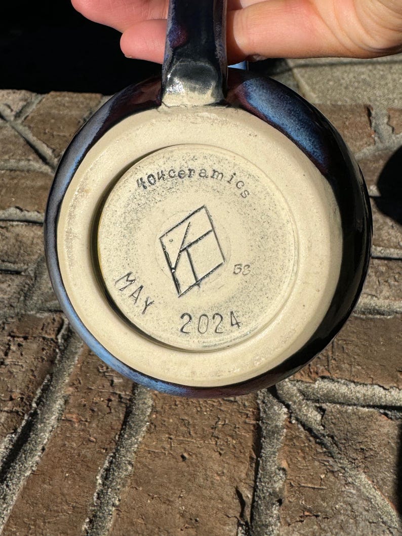 May include: A close-up of the base of a dark blue ceramic mug with a white glaze. The mug has a white stamp that reads "MAY 2024" and "404ceramics" with a logo in the centre.