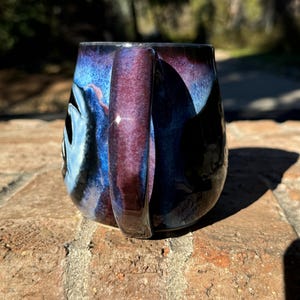 May include: A ceramic mug with a dark blue, purple and black glaze. The mug has a rounded shape and a handle.