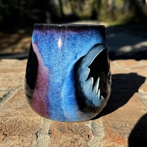 May include: A ceramic mug with a blue, black and purple glaze. The mug has a unique, abstract design.