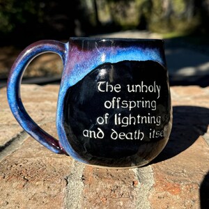 May include: A black ceramic mug with a blue and purple glaze. The mug has a handle and a quote on the front that reads "The unholy offspring of lightning and death itself."