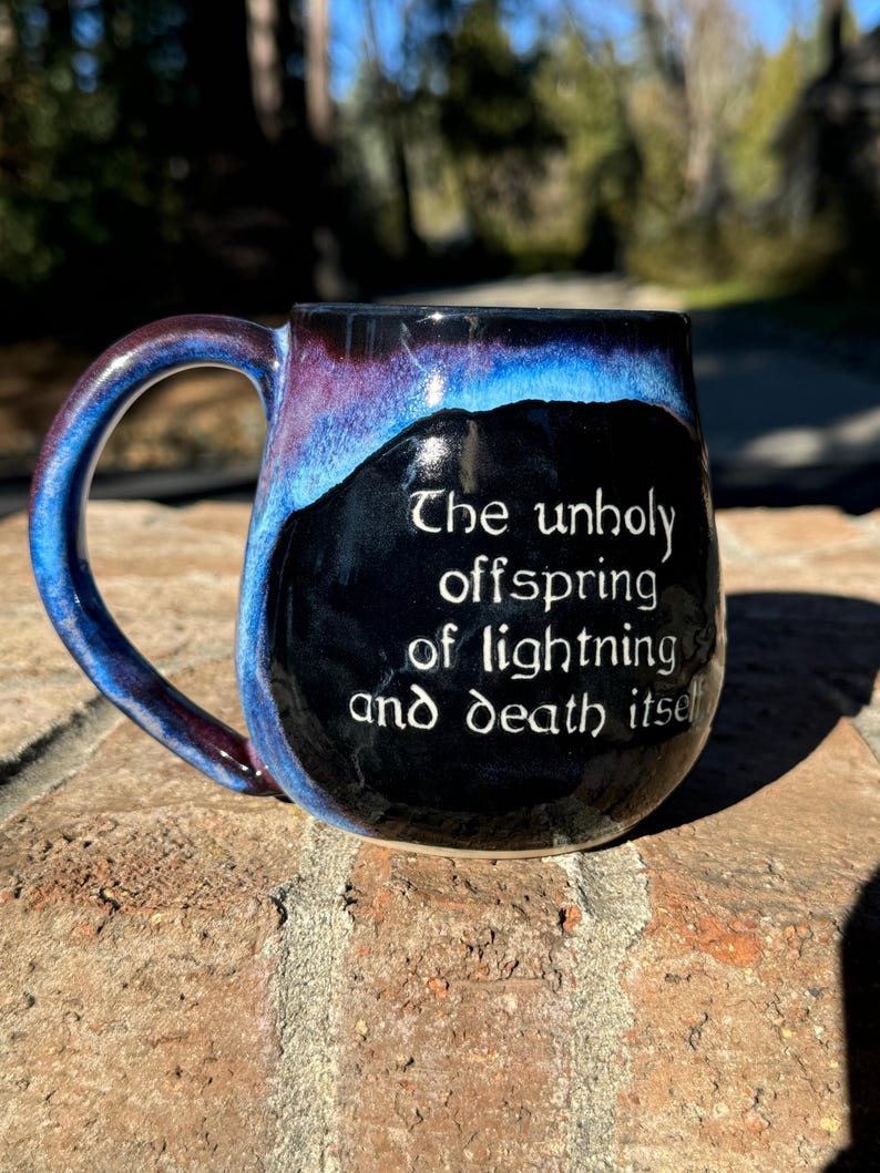May include: A black ceramic mug with a blue and purple glaze. The mug has a handle and a quote on the front that reads "The unholy offspring of lightning and death itself."