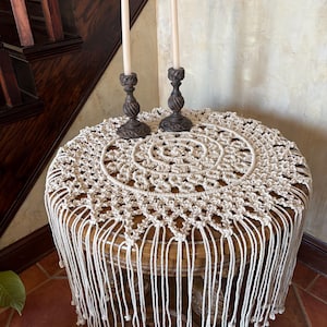 May include: A round wooden table with a cream macrame table cover and fringe. Two dark brown candle holders with white candles are on top. The table is in a room with a wooden staircase.