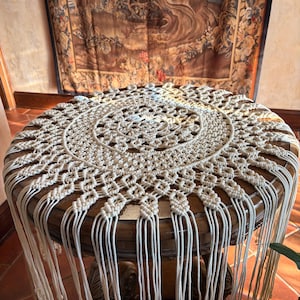 May include: A circular off-white macrame doily featuring an intricate central knot pattern, a radiating geometric design, and long, loose fringe hanging from the outer edge. The textile is crafted with thick cord and displays a symmetrical, knotted structure.