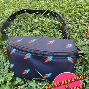 May include: Black fanny pack with a repeating pattern of blue and orange rockets. The bag has a black adjustable strap and a zipper closure. Text on the image reads "Water Resistant & Adjustable."