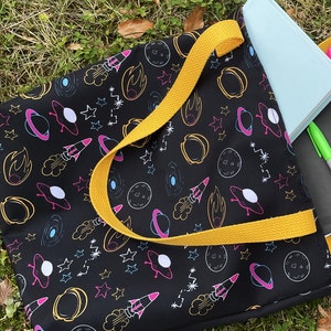 May include: Black tote bag with a yellow strap, featuring a colorful space-themed pattern of planets, rockets, and stars. A light blue notebook and a green marker are visible inside the bag.