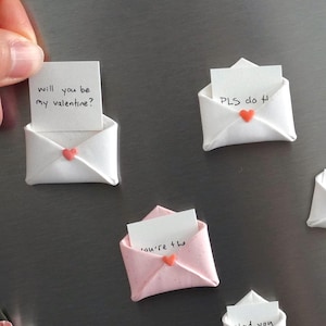 May include: A collection of white and pink origami envelopes, each with a small heart detail and a handwritten message. The notes say "Will you be my valentine?", "PLS do H", "You're the", "I'd Love eve", and "Glad you".