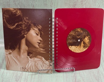May include: A red spiral bound notebook with a vinyl record attached to the cover. The record is red with a gold label that says "Fearless (Taylor's Version)" and features a photo of Taylor Swift. The notebook cover features a sepia toned photo of Taylor Swift.