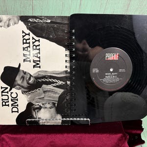 May include: A spiral-bound notebook with a black cover and a vintage vinyl record. The left side features a black and white image of Run DMC and the text "MARY MARY". The record label reads "PROFILE" and "MARY MARY RUN-D.M.C."