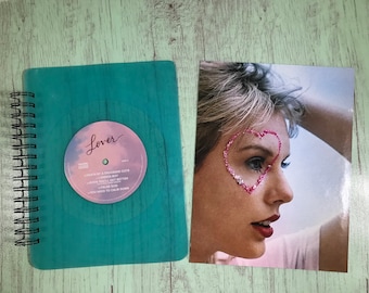May include: A teal spiral-bound notebook with a vinyl record design that says "Lover" and a photo of a woman with a pink heart on her face. The notebook has a wood grain texture.