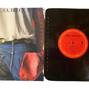 May include: A vintage vinyl record album by Bruce Springsteen, titled "Born in the U.S.A." The album cover features a person in blue jeans and a white shirt. The record label is red with white text.