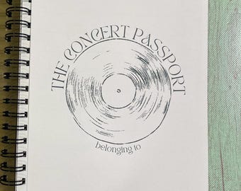 May include: A white notebook with a black spiral binding. The cover features a black and white illustration of a vinyl record with the text "The Concert Passport" and "belonging to".