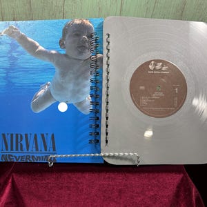 May include: A spiral-bound notebook with a blue cover featuring the Nirvana album "Nevermind" artwork. The notebook is open, revealing a silver-colored page resembling a vinyl record. The record label is visible.
