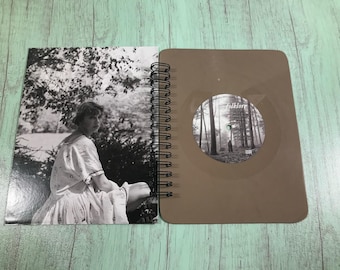 May include: A brown spiral bound notebook with a black and white photo of a woman in a forest on the left side and a brown vinyl record with the text "folklore" on the right side.