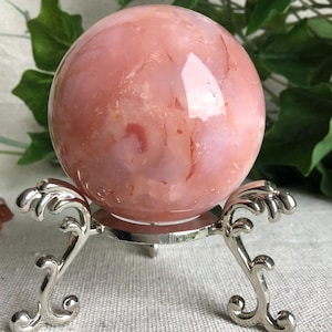 Pode incluir: Uma pedra esférica de cor rosa polida com padrões de redemoinho em tons de coral mais claros e escuros. A esfera repousa sobre um suporte de exibição de metal prateado com pernas curvas ornamentadas e uma base circular de suporte.