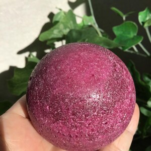 May include: A spherical bath bomb with a deep magenta, textured surface. The object has a porous, granular appearance with subtle crystalline highlights throughout its round form, reflecting light across its dense, dark pink, and slightly uneven exterior.