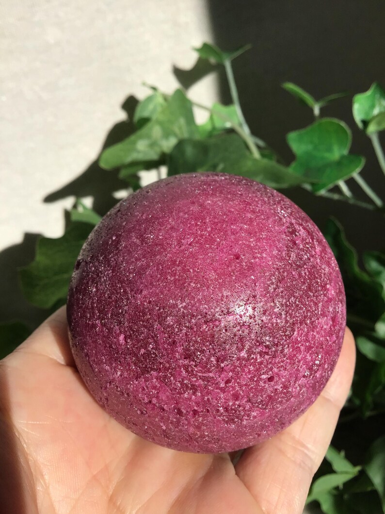 May include: A spherical bath bomb with a deep magenta, textured surface. The object has a porous, granular appearance with subtle crystalline highlights throughout its round form, reflecting light across its dense, dark pink, and slightly uneven exterior.
