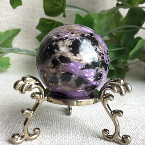 Peut inclure: Une pierre précieuse de charoïte polie et sphérique présentant des motifs tourbillonnants de violet profond, de blanc et d'inclusions minérales noires. La pierre repose sur un support tripode décoratif en métal argenté aux pieds incurvés.