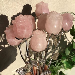 Könnte beinhalten: Ein Strauß aus rosa Rosenquarzblumen mit silberfarbenen Stielen und Blättern. Die Rosenquarzblüten sind in detaillierte Rosenformen geschnitzt. Die Anordnung wird in einer Hand gehalten, mit einem neutralen Hintergrund.