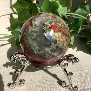 Peut inclure: Une sphère de pierre précieuse polie avec des motifs rouges, gris et beiges, présentée sur un support en métal argenté avec des pieds courbes et ornés. La sphère mesure environ 7.6 cm de diamètre. Des feuilles vertes sont en arrière-plan.