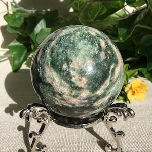 Pode incluir: Uma pedra esférica polida de cor verde com inclusões minerais brancas e castanhas, assente num suporte decorativo de metal prateado de três pernas. A superfície da pedra é lisa e reflexiva, exibindo padrões naturais.