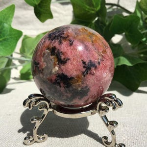 Könnte beinhalten: Eine polierte Rhodonit-Kugel mit einem gesprenkelten Muster aus rosa, schwarzen und goldbraunen Mineralieneinschlüssen. Der Stein ruht auf einem silberfarbenen Metallständer mit drei geschwungenen Beinen und einem runden Ring.