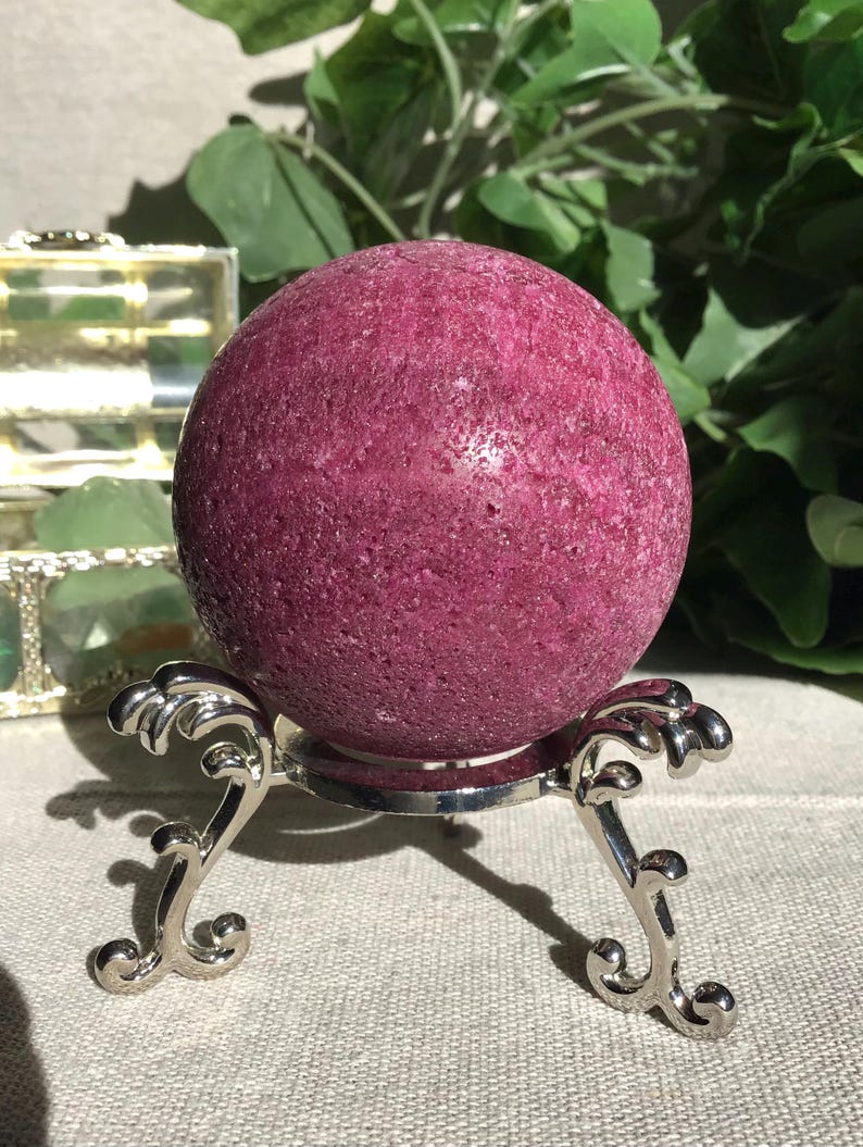May include: A deep magenta, textured spherical stone resting on a polished silver-toned metal display stand with three ornate, curved legs. The surface of the sphere is porous and granular, showcasing a consistent dark pink hue throughout its form.