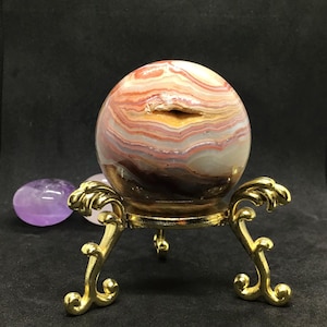 Op de afbeelding: Een gepolijste, bolvormige agaatsteen met horizontale lagen in wit, crème, oranje en donkerbruin met een kleine centrale opening, rustend op een decoratieve goudkleurige metalen driepootstandaard met sierlijke, gebogen poten.