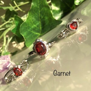 Könnte beinhalten: Drei silberne Ringe mit roten Granat-Edelsteinen. Ein Ring hat einen oval geformten Stein, ein anderer einen herzförmigen Stein und der dritte einen kleineren ovalen Stein. Das Wort "Garnet" ist in schwarzer Schrift geschrieben.