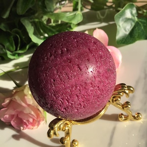 Könnte beinhalten: Eine tiefmagentafarbene Kugel mit strukturierter Oberfläche, präsentiert auf einem goldfarbenen dekorativen Ständer. Der Ständer hat ein verziertes, gerolltes Design. Zarte rosa Blüten und grünes Laub sind im Hintergrund.