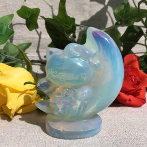 Könnte beinhalten: Eine durchscheinende, schillernde Opalit-Schnitzerei in Form einer Katzenfigur, die auf einem Halbmond sitzt. Die Figur hat eine glatte, polierte Oberfläche, die Licht in Blau- und Weißtönen reflektiert, und steht auf einem runden Sockel.
