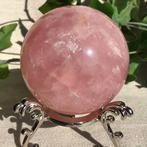 Könnte beinhalten: Eine große, polierte Rosenquarz-Kugel, die auf einem dekorativen, silberfarbenen Metallständer ruht. Die Kugel hat eine zarte rosa Farbe mit natürlichen Variationen. Grünes Laub ist im Hintergrund zu sehen.