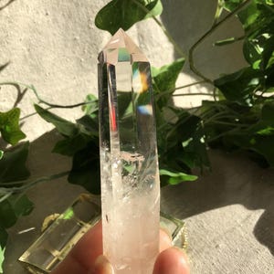 Può includere: Una punta di cristallo di quarzo trasparente a sei lati con una terminazione appuntita. Il cristallo è traslucido, presenta inclusioni interne visibili e un effetto di rifrazione prismatica arcobaleno vicino alla sommità.