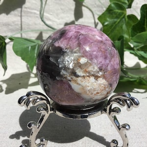 Può includere: Una sfera di lepidolite lucida con sfumature viola, lavanda e grigio scuro con inclusioni minerali bianche. La sfera poggia saldamente su un espositore in metallo color argento con tre gambe curve decorate e una base circolare.