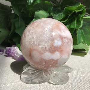 May include: A spherical crystal in shades of pink and white, displayed on a clear, carved glass stand. The sphere has a marbled pattern. Green foliage is in the background, with some purple crystals visible.