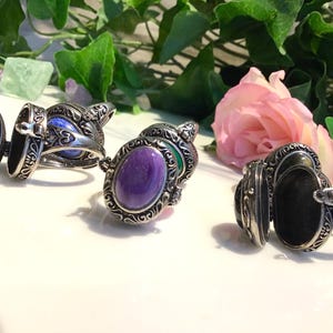 Könnte beinhalten: Silberfarbene Medaillonringe mit ovalen Fächern, einige offen, um blaue, violette und grüne Steine ​​zu zeigen. Die Ringe haben aufwendige, detaillierte Designs und werden mit einer rosa Rose und Grünzeug präsentiert.