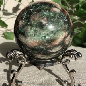 Könnte beinhalten: Ein polierter, kugelförmiger Stein mit einem marmorierten Muster aus tiefem Waldgrün, dunklem Anthrazit und sanften rosa-beigen Farbtönen. Die Kugel ruht auf einem silberfarbenen Metallständer mit drei verzierten, geschwungenen Beinen und einem runden Fuß.