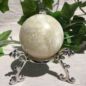 May include: A smooth, off-white, polished stone sphere with subtle internal veining and crystalline textures. The sphere rests securely on a silver-colored metal display stand featuring three curved, ornate legs with scroll-like detailing.
