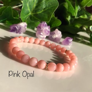 Peut inclure: Bracelet de perles en opale rose avec des détails dorés. Le bracelet est composé de perles rondes rose clair. L'image montre également de petits cristaux d'améthyste violets et des feuilles vertes.