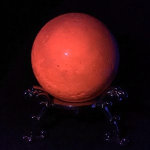 May include: A glowing orange spherical object with a textured surface resting on a dark, metallic, three-legged stand. The sphere emits a bright, warm light against a completely black background, highlighting its cratered, moon-like appearance.