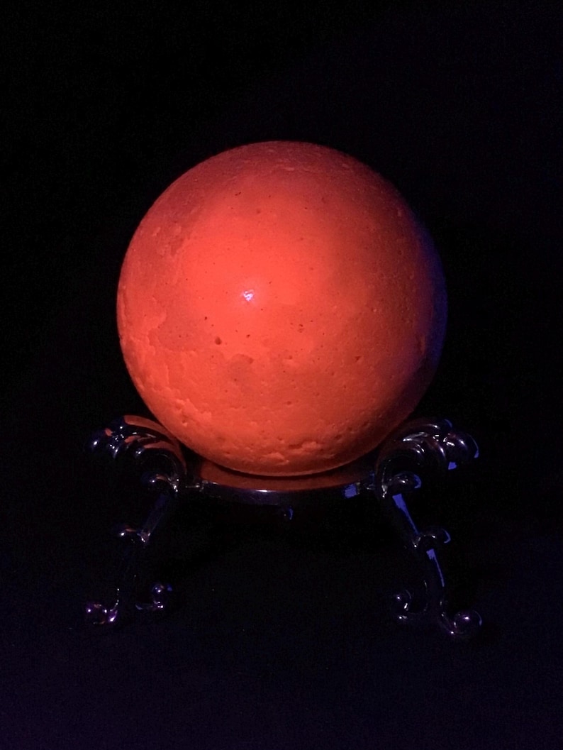 May include: A glowing orange spherical object with a textured surface resting on a dark, metallic, three-legged stand. The sphere emits a bright, warm light against a completely black background, highlighting its cratered, moon-like appearance.