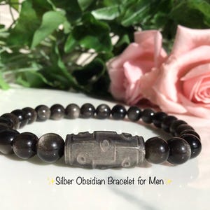 Puede incluir: Una pulsera compuesta por cuentas redondas y pulidas de obsidiana negra y plateada. El centro presenta una cuenta cilíndrica más grande de obsidiana plateada grabada con un patrón geométrico. El texto de abajo dice: Silber Obsidian Bracelet for Men.