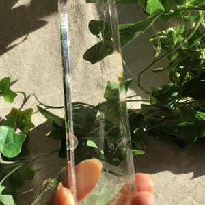 Könnte beinhalten: Ein klarer, transparenter Quarzkristallstab in sechseckiger Form mit einer spitzen Oberseite. Der Kristall hat eine glatte, polierte Oberfläche mit sichtbaren inneren Einschlüssen und natürlichen Brüchen, die das Licht einfangen.