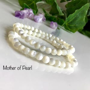 Könnte beinhalten: Zwei Stränge weißer Perlmutt-Perlenarmbänder. Die Perlen sind rund und haben einen glänzenden Glanz. Der Text "Mother of Pearl" steht unten. Amethystkristalle und grüne Blätter im Hintergrund.