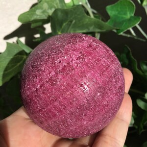 May include: A spherical, deep magenta-colored stone with a rough, crystalline texture. The surface of the sphere displays natural variations in color and mineral composition, featuring a dense, granular appearance with subtle sparkling facets throughout.