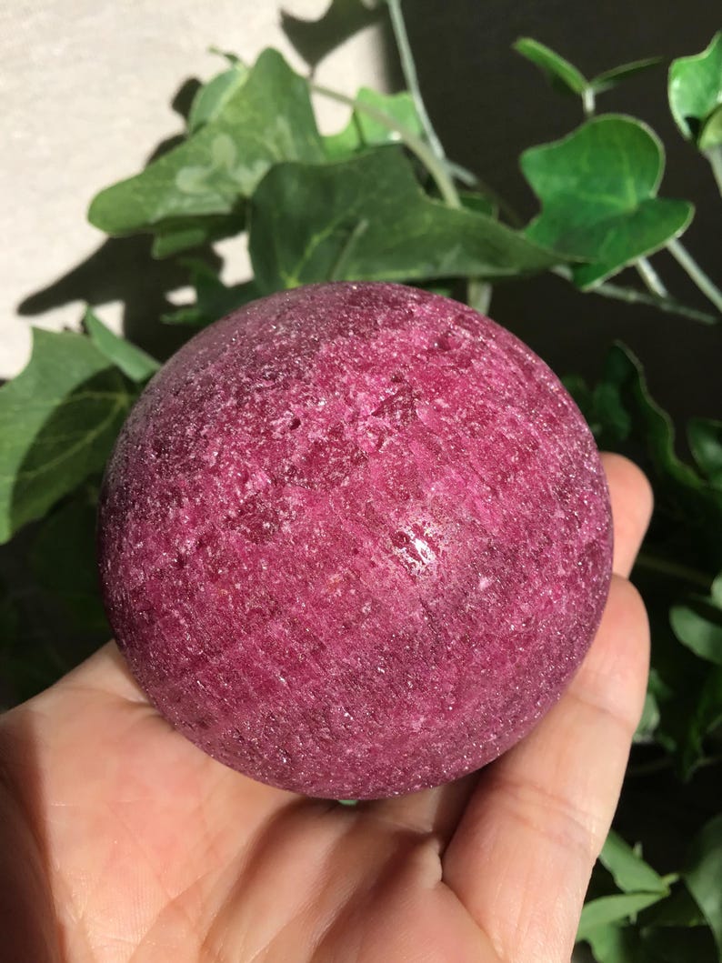 May include: A spherical, deep magenta-colored stone with a rough, crystalline texture. The surface of the sphere displays natural variations in color and mineral composition, featuring a dense, granular appearance with subtle sparkling facets throughout.