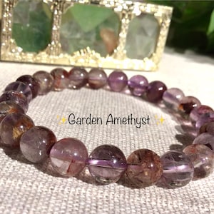 Könnte beinhalten: Ein Armband aus runden, polierten Garten-Amethyst-Perlen mit durchscheinenden violetten Farbtönen und dunklen, erdigen Einschlüssen. Der Text Garden Amethyst ist in der Mitte der kreisförmigen Anordnung der Perlen aufgedruckt.