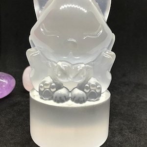 Könnte beinhalten: Eine durchscheinende, hellfarbene Fuchsskulptur. Der Fuchs sitzt auf einem weißen, zylindrischen Sockel, wobei seine Pfoten und seine Brust sichtbar sind. Der Fuchs hält eine Herzform. Die Figur besteht aus einem glatten, glänzenden Material.