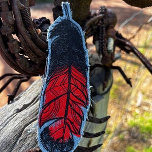 May include: An embroidered fabric patch in the shape of a feather. The top and bottom sections are black, while the centre features a vibrant red colour with detailed black stitching outlining the feather's barbs and central shaft.